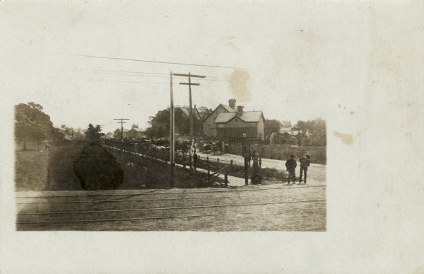 View across the railroad crossing at Hales Corners. Wagons are parked along the side of the road in the background. Two men are standing on the corner near the street sign. Buildings and barns are in the background.
