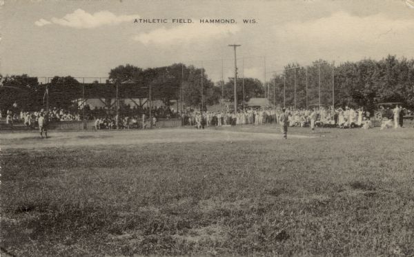 View across an athletic field with a baseball game in progress. Spectators are sitting in the stands and standing behind the base line.