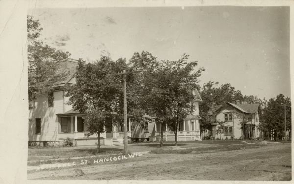 View across unpaved street towards a row of houses. Caption reads: "Residence St., Hancock, Wis."