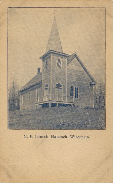 View up slope towards a wooden church with a corner steeple. Caption reads: "M.E. Church, Hancock, Wis."