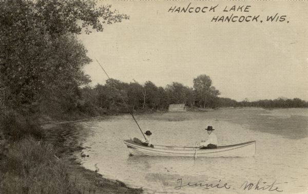 View along shoreline towards two people in a rowboat on a lake. A man is rowing the boat, while a child is fishing is sitting in the back of the boat, fishing with a long pole. There is a boathouse on the shore in the background. Caption reads: "Hancock Lake, Hancock, Wis."