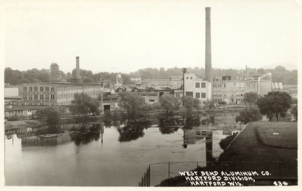 Elevated view across the Rubicon River towards the factory on the far shoreline. Caption reads: "West Bend Aluminum Co., Hartford Division, Hartford, Wis."