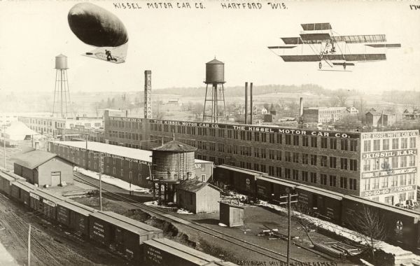 Elevated view of the Kissel auto plant in between the railroad and the river. A dirigible and bi-plane have been cut and pasted onto the image. Caption reads: "Kissel Motor Car Co. Hartford Wis."