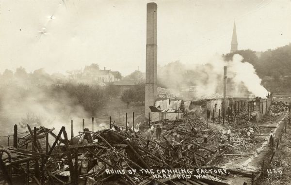 Slightly elevated view of the aftermath of a fire at a canning factory. Men are inspecting the charred ruins, which are smoking. Buildings and a church tower are in the distance. Caption reads: "Ruins of the Canning Factory, Hartford, Wis."