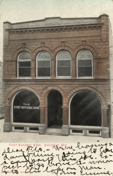 View towards the front of the First National Bank, a red limestone building with large arched windows flanking the arched entrance. A row of four arched windows are on the second floor. Caption reads: "First National Bank, Hartford, Wis."