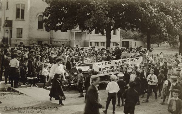 Elevated view of a crowd gathered for the arrival of Buster Brown at the high school. Buster is in the backseat of a roadster.