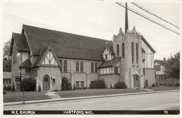 View across street towards the M.E. Church, with Tudor-influenced architecture. Caption reads: "M.E. Church, Hartford, Wis."