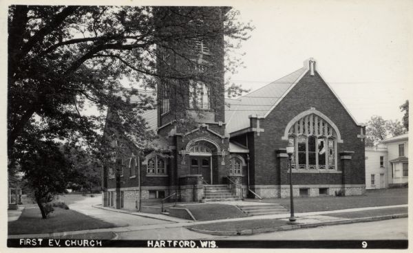 Photographic postcard view across street towards the First Evangelical Church. A lamppost is at the curb in front, and a driveway curves along the left side of the building. Caption reads: "First E.V. Church, Hartford, Wis."