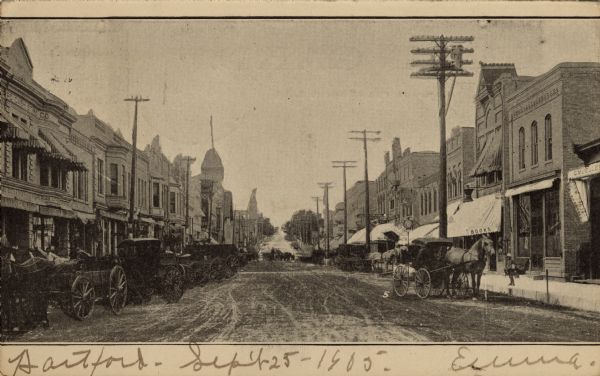 View down center of unpaved, busy city street, with several horse-drawn vehicles parked along the curbs.