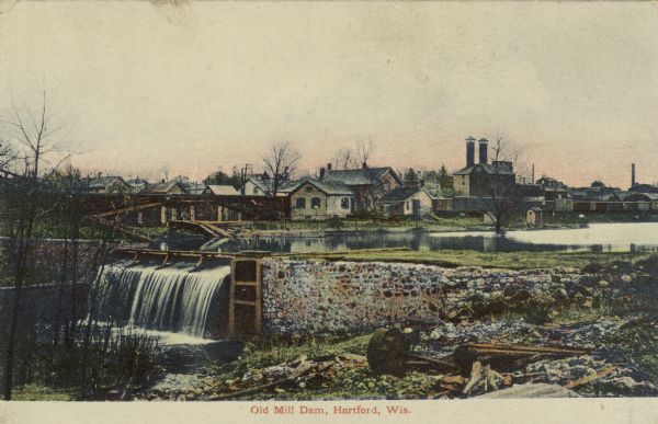 A litho-chrome postcard of a dam on the Rubicon River. There is a railroad yard on the far bank. Caption reads: "Old Mill Dam, Hartford, Wis."