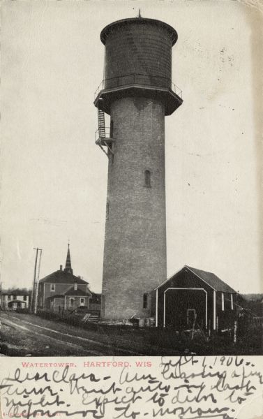 View of the water tower. A stable is to the right at the base of the tower on the right. Other buildings are in the background on the left. Caption reads: "Watertower, Hartford, Wis."