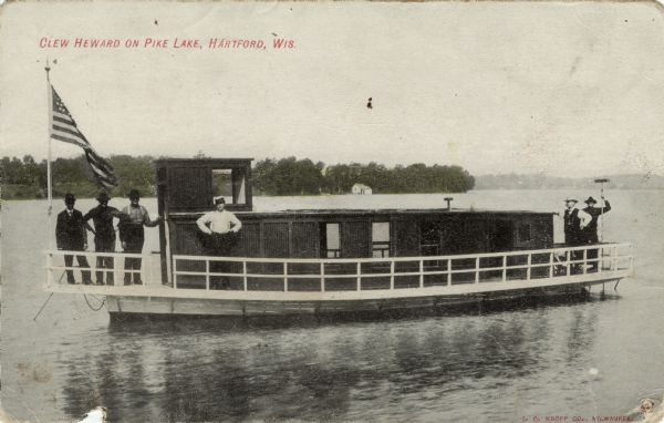 View across water towards the excursion boat, "Clew Heward." A flag is at the bow, and six men are standing on the deck. Caption reads: "Clew Heward on Pike Lake, Hartford, Wis."