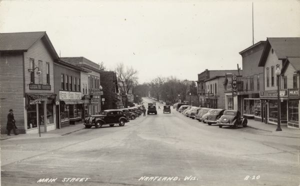View down center of Main Street (part of Hwy 16). Automobiles are parked at an angle along the curbs on both sides. Businesses include two grocers, a drug store, a restaurant, and a Ford dealership. Caption reads: "Main Street; Hartland, Wis."
