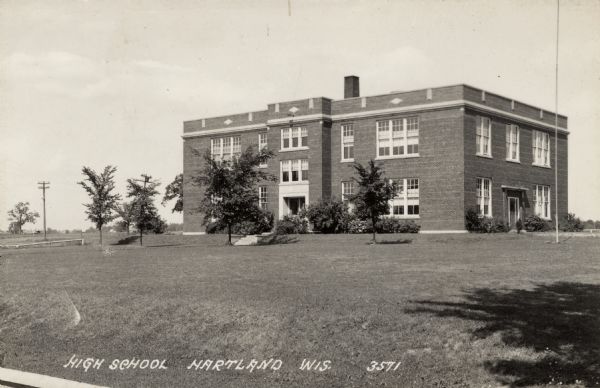View across lawn towards a two-story brick high school. Young trees and shrubs have been planted on the grounds. Caption reads: "High School, Hartland, Wis."