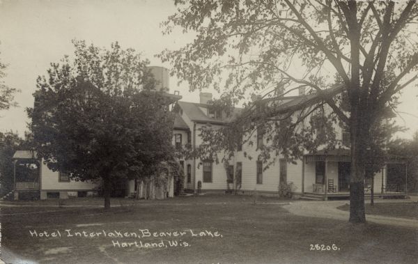 View across lawn towards the Hotel Interlocken. There are chairs on the porch, and a water tower, obscured by a tree, is on a platform just above the roofline. Caption reads: "Hotel Interlaken, Beaver Lake, Hartland, Wis."