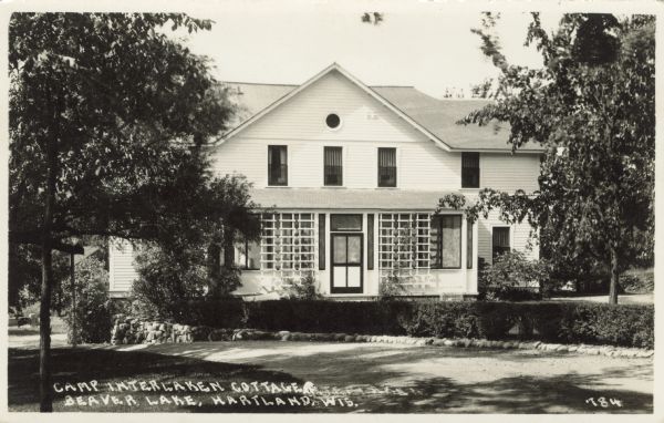 View across driveway towards a guest cottage at the Hotel Interlocken. Trellises are flanking the door on the screened porch. Caption reads: "Camp Interlocken Cottage, Beaver Lake, Hartland, Wis."