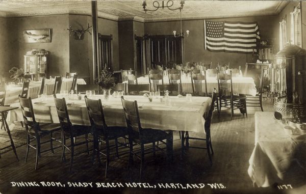 Interior view of the Shady Beach Hotel's dining room. Tables that seat ten are set with drinking glasses. A flag is mounted on the back wall. On the far left is a china cabinet under a mounted taxidermied fish. Antlers are mounted on the wall nearby. Caption reads: "Dining Room, Shady Beach Hotel, Hartland, Wis."