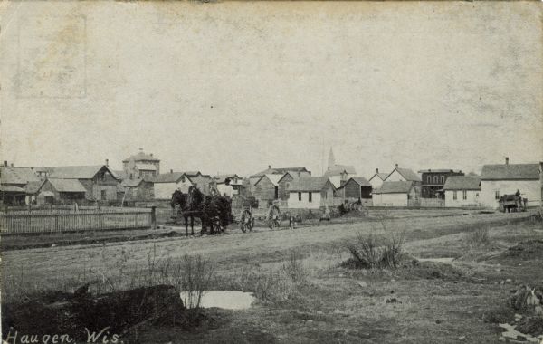 View towards an unpaved road on the edge of Haugen. A man is standing near horses and a wagon on the road. Caption at bottom reads: "Haugen, Wis."