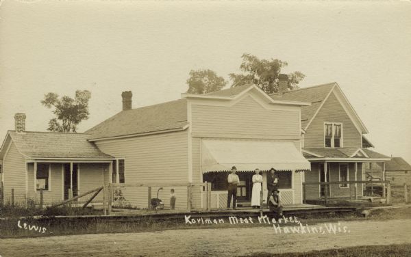 View across unpaved street towards a butcher shop with an awning over the front windows. There is an adjacent house on the right. Four people are posing on the wooden sidewalk in front, and a young child is standing behind them at the corner of the building on the left. Caption reads: "Korlman Meat Market, Hawkins, Wis."