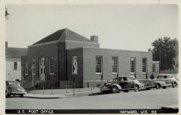 View of a brick post office building. Lampposts at the entrance. Automobiles parked at the curb. Caption reads: "U.S. Post Office, Hayward, Wis."