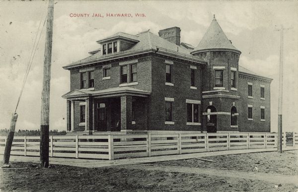 View from street towards the county jail (and likely the county courthouse) surrounded by a fence. Caption reads: "County Jail, Hayward, Wis."