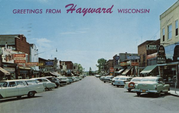 View down Main Street with businesses on both sides, including drug stores, grocery stores, appliance stores and an insurance/real estate office. Automobiles are parked at an angle along the curbs on both sides. Caption reads: "Greetings from Hayward, Wisconsin." Text on back reads: "Muskie Capital of the World. Noted for its Beautiful Lakes, Excellent fishing and resort accommodations. Photo by Bob Young."