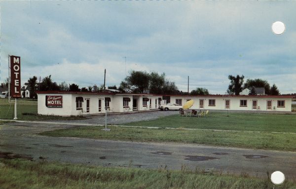 Exterior view of the Lake Hayward Motel. One story L-shaped white building. There is a gravel parking lot, and patio furniture with an umbrella is on the lawn.