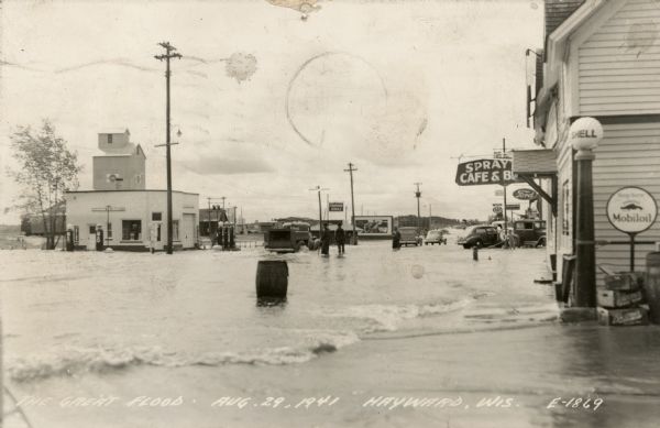 View of a flooded road with a truck and cars making their way through. Men are standing knee-deep in the water. A Standard Oil Station is on the left. There is a Shell and a Mobil Oil sign in the foreground, and a Ford dealership is further down the street on the right.