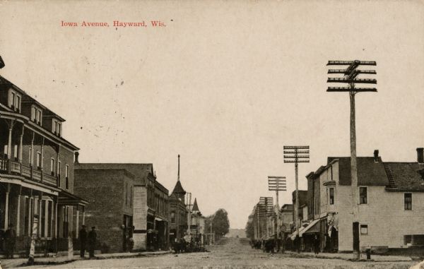 View down a commercial street lined with businesses. A barber pole is on the left, and a fire hydrant is on the right street corner. Large power poles are on the right side of the street. Caption reads: "Iowa Avenue, Hayward, Wis."