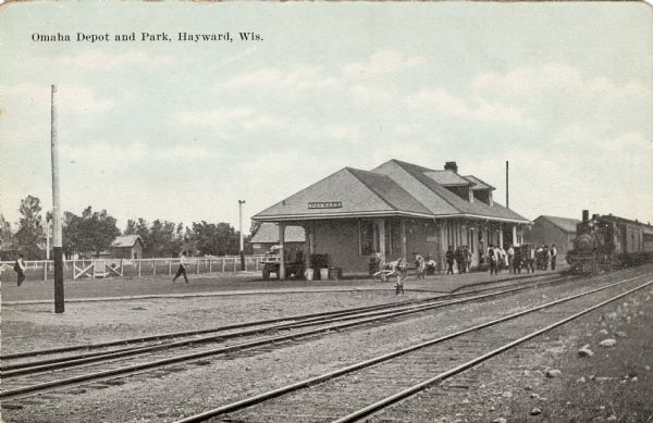 View across railroad tracks towards the Hayward Depot, with a train approaching. Several people are on the platform.