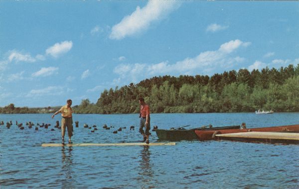 Two men are log rolling on the Namakagon River at the North Wisconsin Logging Camp and Museum. Rowboats are moored next to a dock.