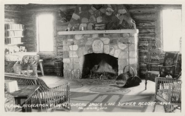 View of a sitting room in the Le Toureau Spider Lake Summer Resort recreation building. A stuffed bobcat is on the mantle. Shelves of books are above a deskon the left. Chairs are around the stone fireplace.