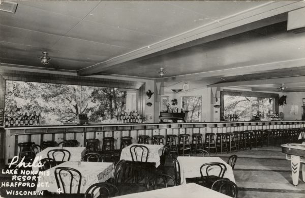 Kodak photographic postcard of the interior of a restaurant with a long bar. Bottles are stacked next the picture windows behind the bar. A piano is in an alcove in the background. Caption reads: "Phil's Lake Nokomis Resort, Heafford Jct., Wisconsin."