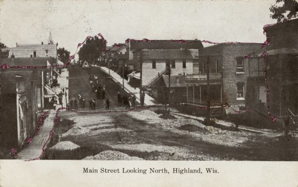 View down hill towards small town street with a dip at the bottom where men are standing, with a group of people on the hill in the background. Caption reads: "Main Street Looking North, Highland, Wis."
