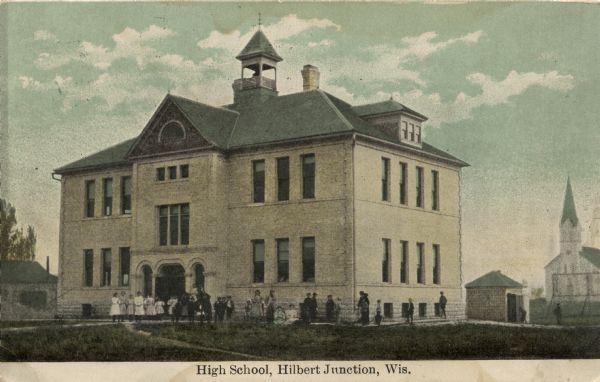 Three-quarter view of the front and side of a brick high school. There are a few students gathered in front. There is a church building in the background on the right. Caption reads: "High School, Hilbert Junction, Wis."