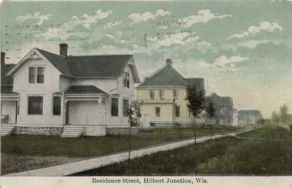 View of a street or sidewalk in front of two-story dwellings. Small trees are planted along the sidewalk. Caption reads: "Residence Street, Hilbert Junction, Wis."