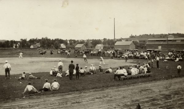 Photographic postcard view of a small town baseball game in progress. Audience members are standing on the sideline and sitting on the grass. Horses and buggies and automobiles are parked on the opposite side of the field.