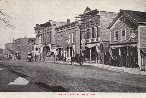 View across street towards businesses. Horse-drawn vehicles are moving down the street, and a group of men are standing in front of a shop on the right. Large power poles are along the sidewalk. Caption reads: "Water Street, Hillsboro, Wis."