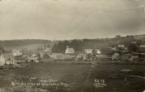 Slightly elevated view of Hillsdale. Two churches are on the edge of the town in the distance, and a field and dwellings are in the foreground. Caption reads: "Bird's [sic] Eye View of Hillsdale, Wis."