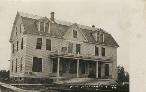 View of a hotel in Holcombe with steps up to a porch with columns at the entrance, and a balcony above. A sign on the side of the building reads: "The Journal." A man is standing at the back corner on the left side of the building. Caption reads: "Hotel, Holcombe, Wis."