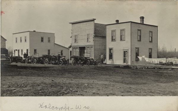View across unpaved street in Holcombe. There is a long row of parked automobiles in front of a barbershop at the end of the block. Laundry is drying on lines on the right. Men are standing on the wooden sidewalk.