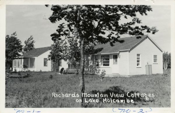 View across tall grass towards two guest cottages at Richard's Mountain View. Caption reads: "Richards Mountain View Cottages on Lake Holcombe."
