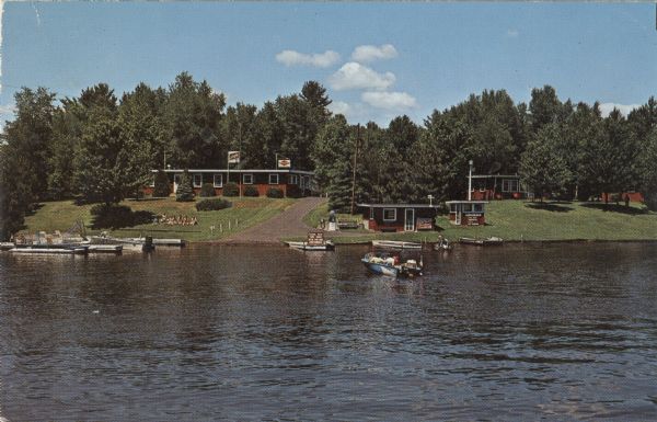 View across water towards a lakefront resort and boat landing. There are people in a few boats in the lake, and other boats tied up at piers. There is tavern at the top of the drive.<p>Text on reverse reads: "Pine Drive Resort Lake Holcombe's Finest Off Highway 27 - 3 miles west on County Trunk M; then 1/4 mile north. R. Rt. 2 Holcombe, Wisconsin 54745 Don & Edna Clark Phone 715-595-4201."</p>