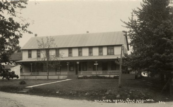Photographic postcard view of a wood frame hotel with a long porch. Caption reads: "Hillside Hotel, Holy Hill, Wis. B. Heileman, Prop."