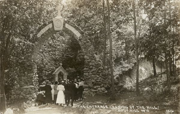 Photographic postcard view of the stone arch marking the entrance to the Stations of the Cross at Holy Hill. A crowd of pilgrims are standing underneath. Caption reads: "The Entrance, Leading Up the Hill, Holy Hill, Wis."