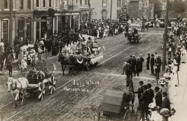 Elevated view of horses pulling floats in the Fourth of July parade down Main Street. Crowds are watching from the sidewalks. Caption reads: "July 4th 08, Horicon, Wis."