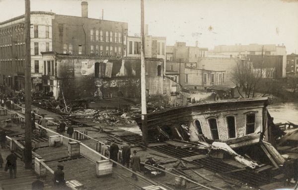 Elevated view of a completely burned building in downtown Horicon at the river. A cigar store is next door, and men are gathered on the bridge.