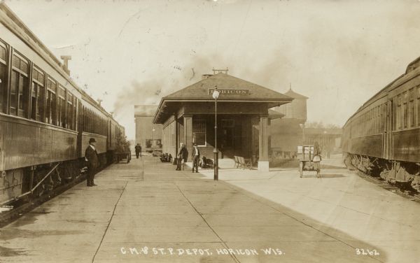 View down platform towards a C. M. & St. Paul train at the depot. A few men are standing on the platform. There is a water tower just behind the depot, and another train is on the railroad tracks on the right. Caption reads: "C. M. & St. P. Depot, Horicon, Wis."