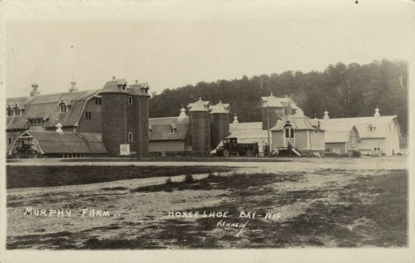 View of a farm with multiple barns and silos. Two trucks are parked on the property.