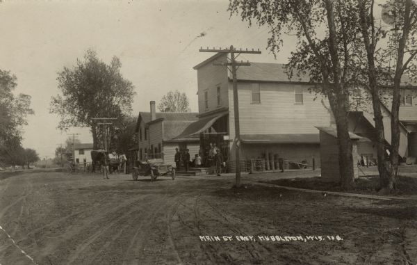 View of Main Street with a general store in the center. An automobile and a horse-drawn vehicle in the street. People are gathered on the front stoop of the store. Caption reads: "Main St. East, Hubbleton, Wis."